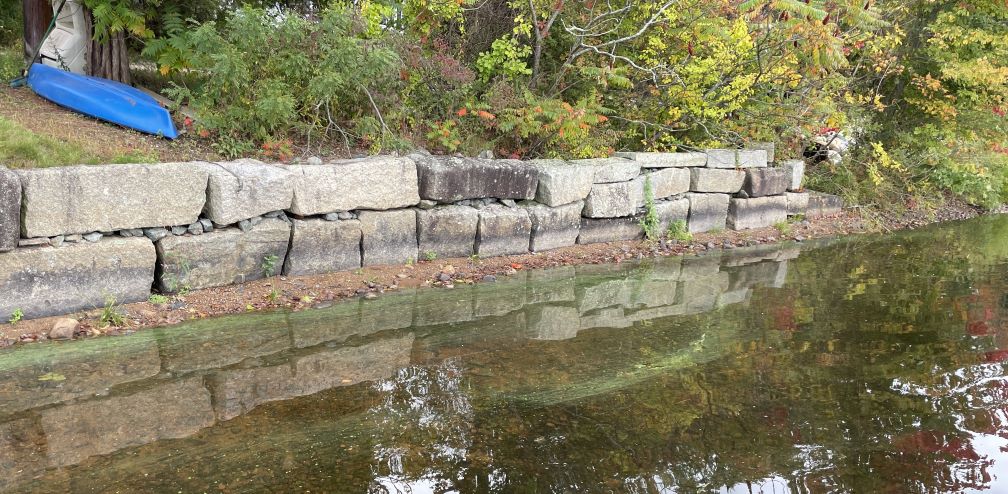 A retaining wall made of a large rectangular gray granite stones. Below the wall is a little bit of sandy/muddy/rocky shore. Calm water comes up to that shore, and there are streaks of green slime paralleling the shore. Above and behind the wall is some shrubbery, and a little grass with an upside down blue kayak.
