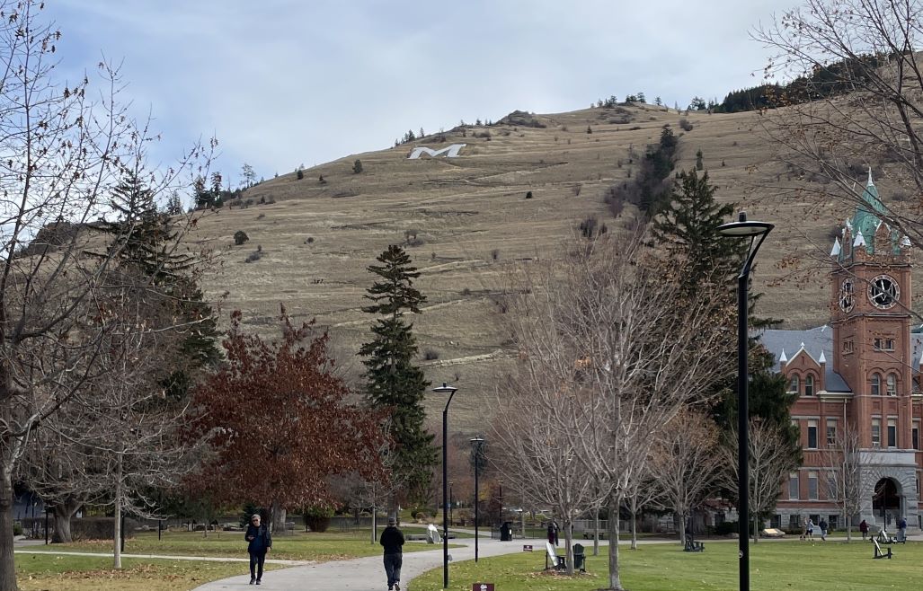In the foreground, a few people are walking on a cement path between trees and lawn. On the right is an old brick building with a clock tower. Directly ahead is a brown grassy hillside with a few trees on it, and a large white capital letter M. There are switchbacks going up the hillside with a number of people walking on them.