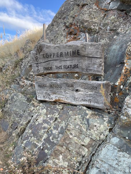 An old weathered sign with words chiseled into wooden boards which says '1890 Copper mine. Enjoy this feature. Please respect our history. Leave as you found it.' On lichen covered rocks.