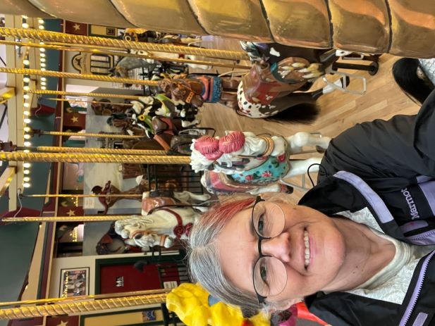 a white woman wearing glasses and a purple and black jacket smiles at the camera. Her hair is salt and pepper and the right-hand side is partially streaked with maroon dye. In the background there are carousel horses without riders. Horses have gold posts through their centers and are in various galloping positions, neck stretched out or bent, legs together or stretching out, horses in all colors that horses come in (white through various brown colors, and black), with fancy saddles and bridles.